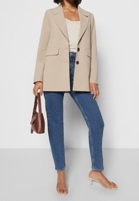 Beige tailored blazer with notched lapels, two front pockets, single button closure, paired with blue high-waisted jeans and clear heeled sandals.