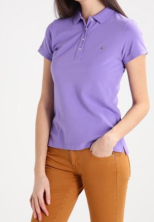 Polo shirt viola con una texture morbida, maniche corte e colletto con tre bottoni; presenta un piccolo logo sul petto. Abbinato a pantaloni arancioni.