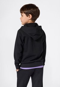 Felpa nera con cappuccio e texture liscia, caratterizzata da una tasca a marsupio e polsini a coste, indossata sopra una maglietta viola.