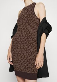 Robe en tricot marron avec un motif géométrique, sans manches, et un ourlet côtelé. Associée à une veste noire tombant sur une épaule.