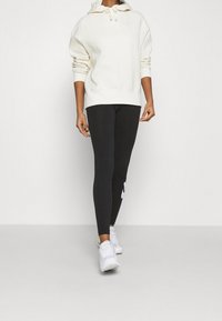 Ljusbeige hoodie med ficka fram och dragsko i huvan, tillsammans med svarta leggings med en stor vit logo. Vita träningsskor.