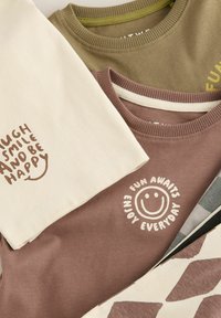 Drie opgestapelde t-shirts in aardetinten met speelse teksten en vrolijke afbeeldingen, die een blijmoedige, ontspannen sfeer uitstralen.