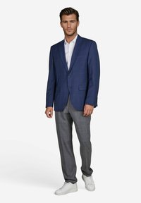 Marineblaues Blazer im Slim Fit, kombiniert mit hellgrauen, strukturierten Hosen und weißen Sneakers. Das Blazer hat zwei Vorderaschen und einen Reverskragen.