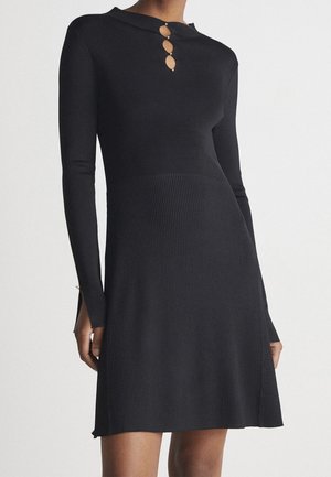 Robe noire en tricot côtelé manches longues avec trois découpes en forme de trou de serrure et boutons en perles au col haut, cintrée à la taille, évasée à la jupe.