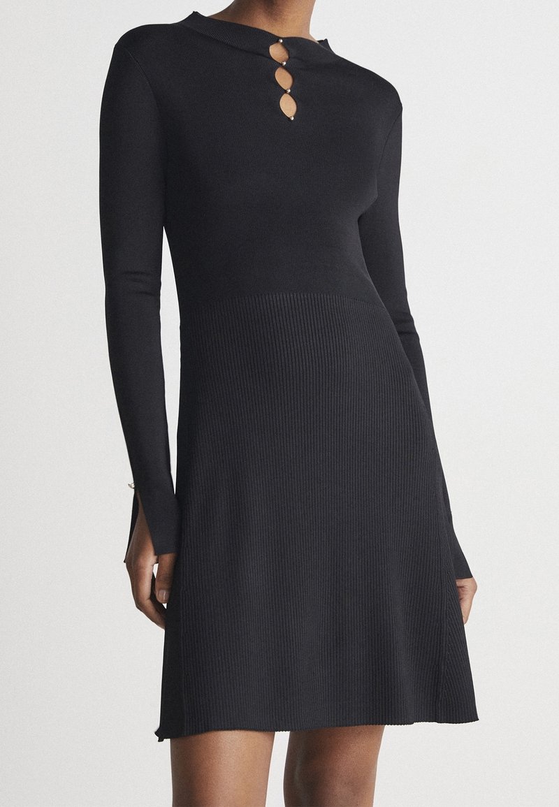 Robe noire en tricot côtelé manches longues avec trois découpes en forme de trou de serrure et boutons en perles au col haut, cintrée à la taille, évasée à la jupe.