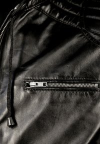 Sorte læderbukser med en lynlårs lomme, snorelukning og struktureret overflade. Lynlåsen har en sølvfinish med et lille træk.