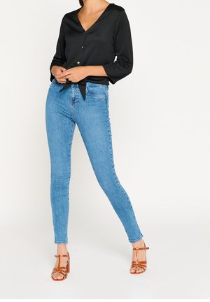 Vrouw draagt lichtblauwe skinny jeans, zwarte blouse met knopen en strik aan de voorkant, en bruine sandalen met hak en bandjes, staand tegen een witte achtergrond.