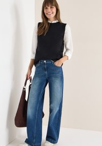 Czarny, bezrękawnik dzianinowy na białej koszuli z kołnierzykiem, zestawiony z szerokimi niebieskimi dżinsami. Brązowa torba na ramię. Neutralne tło.