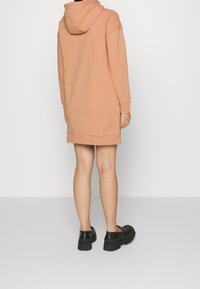 Vestido sudadera color melocotón con capucha, de mangas largas, puños acanalados y corte relajado, que termina por encima de la rodilla. Unos zapatos negros de suela gruesa complementan el conjunto.