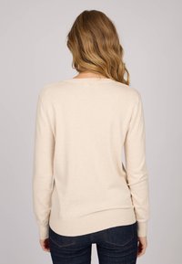 Pull en mailles beige clair avec des manches longues, un col rond et un ourlet côtelé, présentant une texture douce et un design simple.