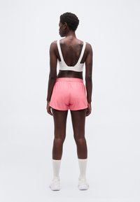 Γυναίκα που στέκεται με την πλάτη γυρισμένη, φορώντας λευκό αθλητικό σουτιέν, ροζ σορτς, λευκές κάλτσες και λευκά αθλητικά παπούτσια σε απλό φόντο.