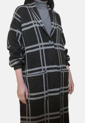 Femme portant un manteau long à carreaux gris foncé avec un bouton unique sur un pull à col roulé gris clair.