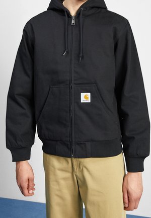 Man draagt een zwart vest met ritssluiting, capuchon en voorzakken, en een beige broek, staand tegen een effen lichte achtergrond.