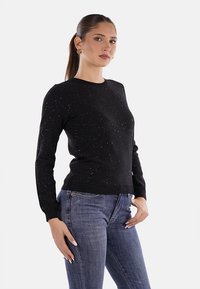 Donna con una lunga coda di cavallo che indossa orecchini a cerchio dorati, un maglione a pois neri e jeans blu, in posa con una mano sulla vita.