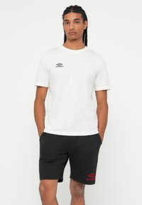 Muškarac s pletenom kosom nosi bijelu Umbro majicu i crne Umbro kratke hlače, stoji s rukama u džepovima na bijeloj pozadini.