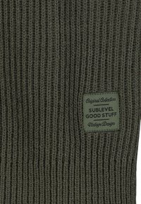 Rippender olivgrüner Strickstoff mit einem eingewebten Etikett, auf dem in schwarzer und weißer Schrift "Sublevel Good Stuff Vintage Design" steht.