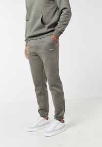 Άτομο που φοράει ελαφρώς πράσινη φόρμα και αντίστοιχο φούτερ με τις χέρια στις τσέπες, συνδυασμένα με λευκά αθλητικά παπούτσια χαμηλού προφίλ.