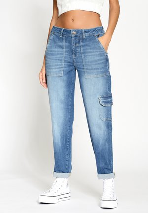Frau trägt hellblaue Denim-Cargohosen mit aufgekrempelten Säumen, ein weißes kurzes Stricktop und weiße High-Top-Sneaker, stehend vor einem einfarbigen Hintergrund.