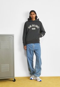 Sudadera gris con "New Balance 1906" impreso en crema, combinada con pantalones vaqueros de pierna recta en azul claro y zapatillas blancas con acentos azules.