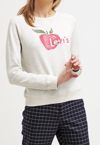 Světle šedá mikina s červeným jablkem a nápisem "Levi's". Má kulatý výstřih a žebrované manžety, doplněná tmavě modrými kostkovanými kalhotami.