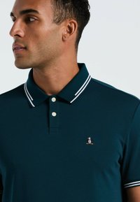 Donkerblauw poloshirt met een witte gestreepte kraag, twee knopen en een klein pinguïnlogo op de borst. Zacht materiaal met een gladde textuur.