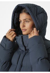 Tmavě modrá péřová bunda s vysokým límcem a velkou kapucí. Disponuje prošívaným designem a odolným materiálem, který poskytuje izolaci a teplo.