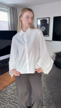 Kvinne med rett blondt hår står innendørs iført en løs hvit skjorte med knapper og grå bukser i en moderne stue.