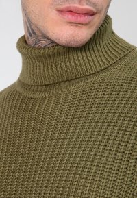 Maglione a collo alto lavorato a maglia di colore verde oliva, caratterizzato da una texture a costine e una silhouette aderente. In primo piano, si notano i dettagli delle cuciture e del design del colletto.