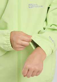 Giacca impermeabile verde chiaro con polsini regolabili. Presenta il logo "Jack Wolfskin" e "TEXAPORE" stampati sulla manica. Chiusura con zip davanti.