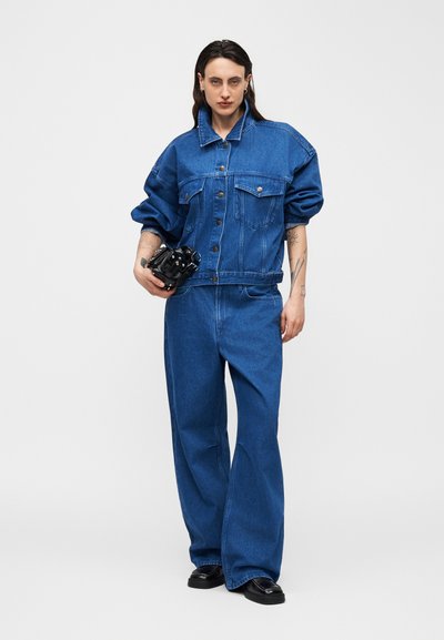 Άτομο που φοράει φαρδιά μπλε τζιν μπουφάν και τζιν παντελόνι με φαρδιά μπατζάκια, κρατώντας μια μαύρη κάμερα σε απλό ανοιχτό φόντο.