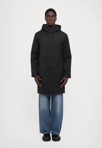 Cappotto lungo nero con cappuccio, dotato di una chiusura a zip, maniche lunghe e taglio dritto. Abbinato a jeans di denim blu larghi e scarpe nere.
