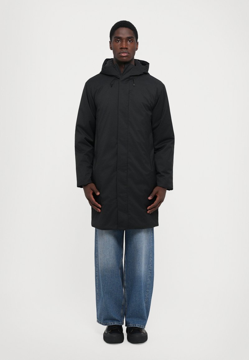 Cappotto lungo nero con cappuccio, dotato di una chiusura a zip, maniche lunghe e taglio dritto. Abbinato a jeans di denim blu larghi e scarpe nere.