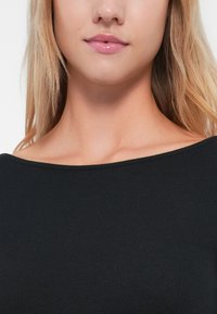 Svart ribbestrikket topp med vid halsutringning, glatt tekstur og tettsittende design som fremhever kragebeinet og skulderområdet.