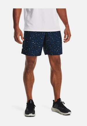 Mand iført marineblå atletiske shorts med mønster, sorte løbesko og en hvid t-shirt, stående mod en hvid baggrund.