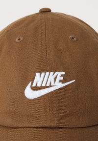 Boné de beisebol de algodão castanho com um logótipo Nike bordado em branco na frente. O tecido tem uma textura ribaltada subtil.