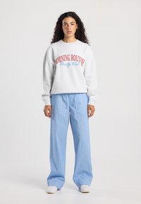 Grijze sweatshirt met "MORNING ROUTE Varsity Club" gedrukt in rood en blauw, gecombineerd met blauwe verticaal gestreepte broek en witte schoenen.