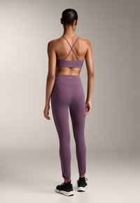 Vijolična športna nedrčka z križnimi naramnicami in ujemajočimi se visokimi hlačami. Temni čevlji z belimi podplati, gladek material, prilegajoča zasnova.