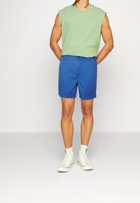 Camisa sin mangas verde, pantalones cortos azules y zapatillas altas blancas. El conjunto presenta un ajuste relajado y un diseño simple con colores sólidos.