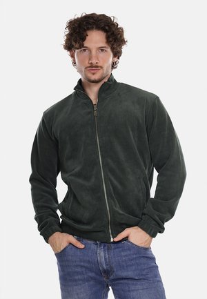 Giacca in fleece verde scuro con colletto alto e zip completa, caratterizzata da una trama morbida e maniche lunghe. Indossata sopra un paio di jeans blu.