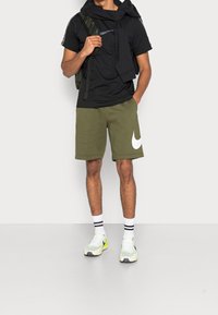 Schwarzes T-Shirt, olivgrüne Sweathorts mit großem weißen Nike-Logo, weiß-gelbe Sneakers, schwarzer Rucksack, gestreifte weiße Socken.