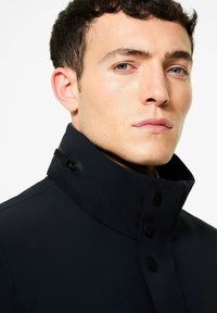 Cappotto blu navy con colletto alto, caratterizzato da bottoni neri e una zip sul colletto. Il tessuto liscio e strutturato mette in evidenza il design moderno.