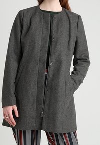 Abrigo de mezcla de lana color gris oscuro con cuello redondo, cierre frontal con botones a presión, bolsillos laterales y corte entallado, presentado sobre pantalones con estampado.