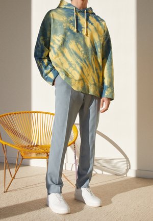 Personne portant un sweat à capuche tie-dye jaune et vert, un pantalon bleu clair et des baskets blanches, debout à côté d'une chaise tissée jaune au soleil.