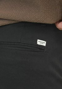 Pantaloni neri con una texture liscia, caratterizzati da un design elegante e una tasca piatta visibile. Un'etichetta con il marchio è attaccata vicino alla tasca.