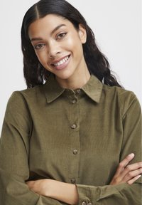 Camicia in corduroy verde oliva con colletto pieghevole, apertura frontale con bottoni e maniche lunghe arricciate, che mette in mostra un tessuto testurizzato e bottoni marroni.