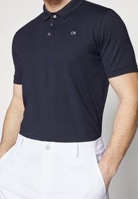 Mörkblå pikétröja med tvåknappsknäppning, korta ärmar och CK-logotyp broderad i vitt. Matchas med vita byxor. Slät textur.