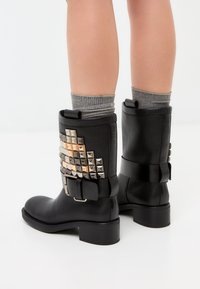 Botas de tobillo de cuero negro con un tacón robusto, adornadas con tachuelas en forma de pirámide en plata y oro a los lados y una amplia correa con hebilla negra.