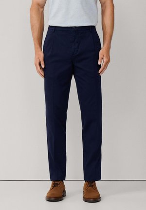 Homme portant un pantalon ajusté bleu marine, une chemise grise rentrée et des chaussures en suède brun à lacets, se tenant contre un fond clair uni.