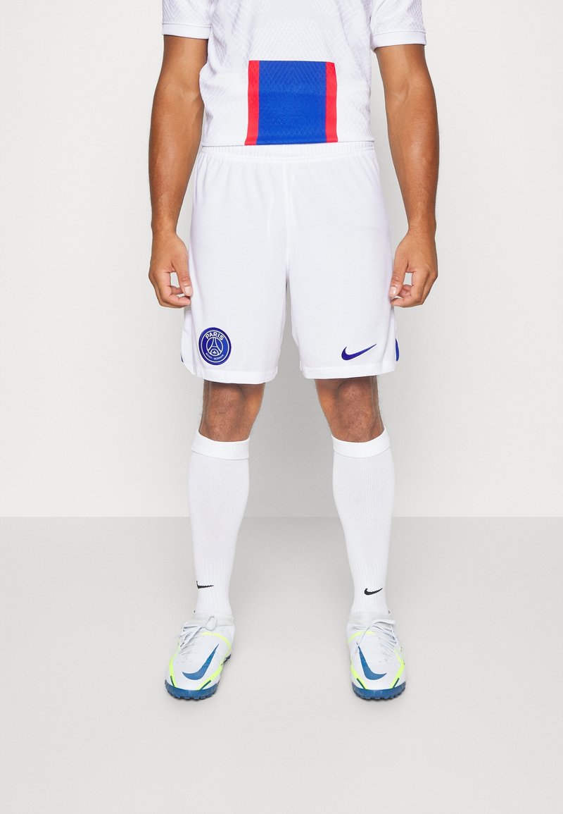 Homme portant un short de football blanc Paris Saint-Germain, des chaussettes blanches et des crampons blancs Nike, debout sur un fond uni.