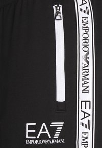 Svarta sportbyxor med en vit dragkedja och vertikal vit branding. Har elastisk midja och mjukt, texturerat tyg. Dragkedjeficka detaljer.